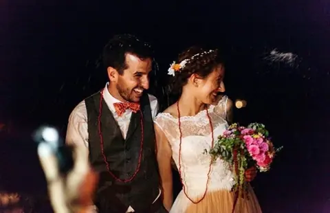 Una pareja recién casada sonríe bajo la noche, con corona de flores y el lazo rojo de la ceremonia de unión