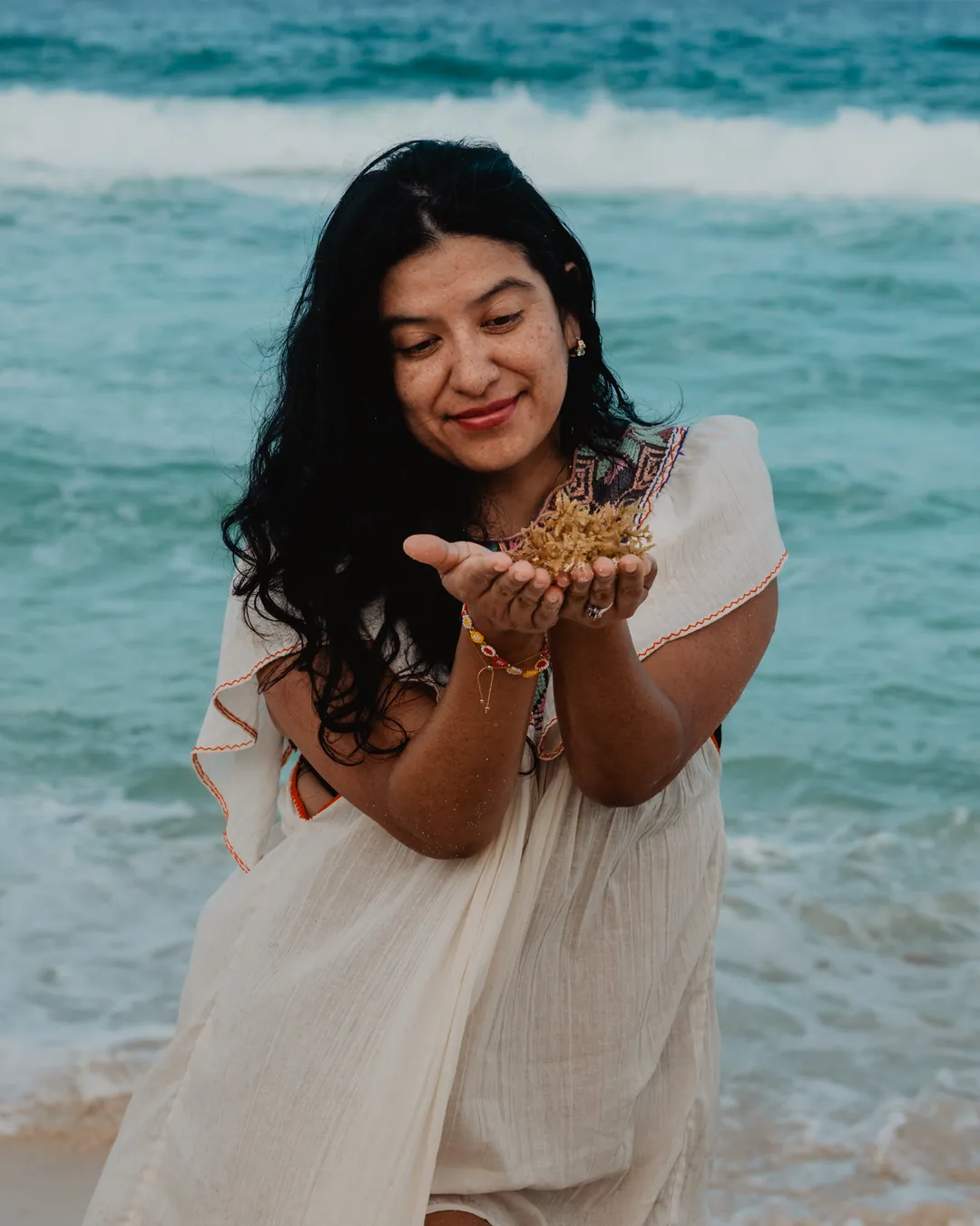 Blanca Coutiño sostiene en el cuenco de las manos una ofrenda dorada del mar, vestido de lino con bordado, frente a las olas turquesa del Caribe